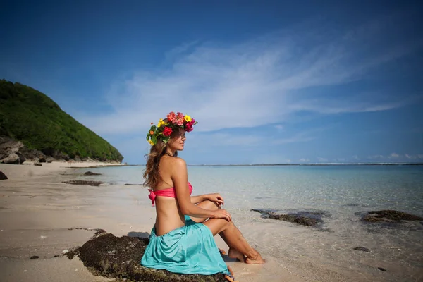 Beautiful girl with flower wreath on her hair is sitting on a beach — Stock Photo, Image