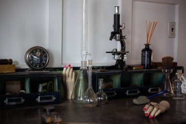 Masanın üzerinde antika şeyler var. Cam mataralar ve test tüpleri, retro mikroskop, oklu eski yuvarlak çalar saat, manken eller.
