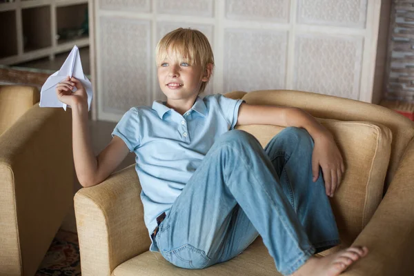 Mavi tişörtlü yakışıklı sarışın adam kanepeye oturup elinde kağıt bir uçak tutuyor ve fırlatmak üzere.