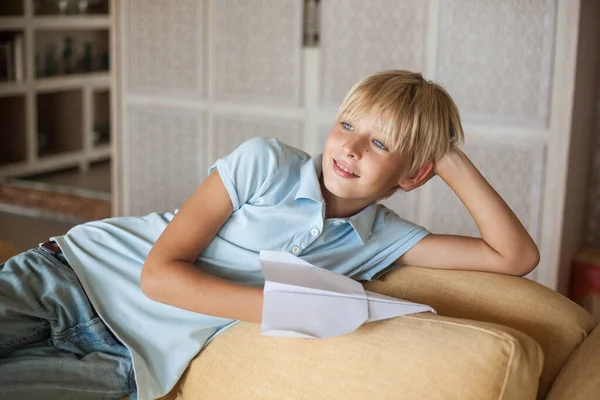 Mavi tişörtlü yakışıklı sarışın adam kanepeye uzanmış ve elinde kağıt bir uçak tutuyor. Oğlan hayallere dalmış.