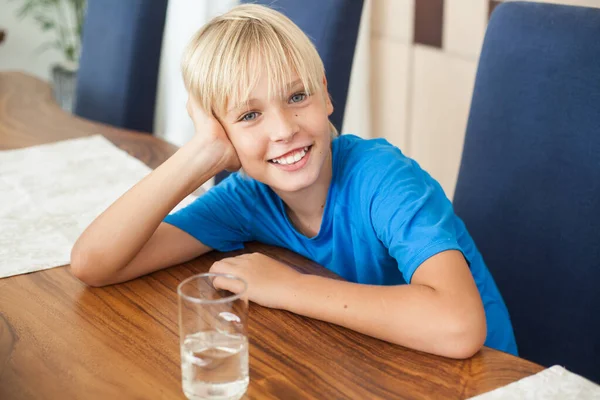 handsome smiling blond teenager guy sitting in a room at a wooden table with a glass of clean drinking water. boy looks at the camera
