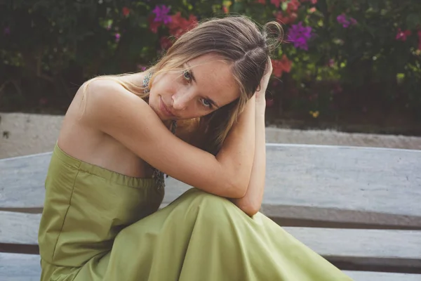 Retrato de una hermosa mujer rubia joven sin hombro vestido largo y verde claro posando en un banco blanco en el jardín. sus codos descansan sobre sus rodillas. tiro al aire libre en el parque durante el día de verano — Foto de Stock