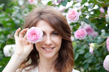 Arka planda pembe gül çalısı olan çekici, gülümseyen esmer kadın portresi. Kız kameraya bakar ve elinde çiçekle bir gözünü kapatır.