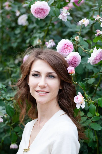 Primer plano retrato de hermosa joven morena con rosa rosal en el fondo. chica mira a la distancia y sonriendo . —  Fotos de Stock