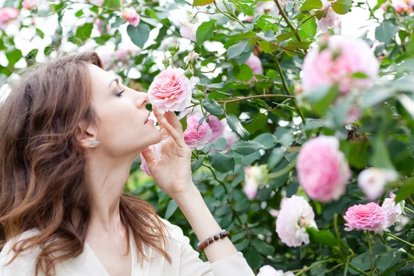 Primer plano retrato de hermosa joven morena con rosa rosal en el fondo. chica se para de perfil y busca una flor y la huele — Foto de Stock