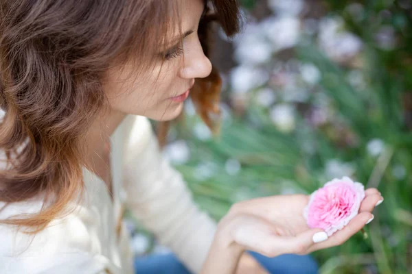 Portrait de profil d'une belle jeune femme brune tenant une fleur rose dans sa main et la regardant. Une fille aux cheveux longs et un chemisier blanc — Photo