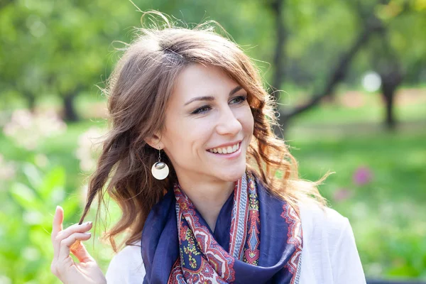 Hermosa joven morena sonriente mujer en un jersey blanco y con una bufanda azul alrededor de su cuello en un parque verde de primavera —  Fotos de Stock