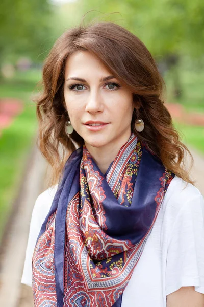 Portrait vertical de belle jeune femme brune en pull blanc et avec une écharpe bleue autour du cou dans un parc vert printanier — Photo