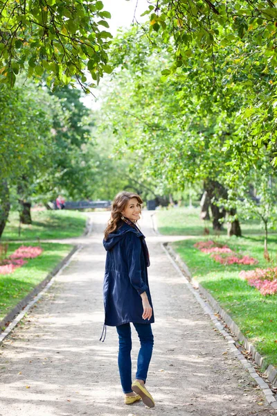 Hermosa mujer morena joven en una chaqueta azul y pantalones vaqueros caminando en un parque verde primavera. Callejón con árboles y flores están en el fondo — Foto de Stock