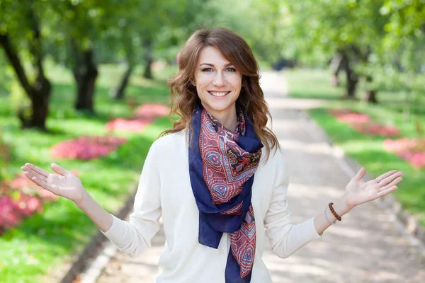 Hermosa joven morena sonriente mujer feliz en jersey blanco y con una bufanda azul en un parque verde primavera. callejón con árboles y flores están en el fondo. chicas manos en una posición de bienvenida abierta — Foto de Stock