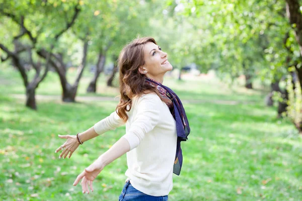 Beautiful young brunette smiling happy woman in white jumper and with a blue scarf in a spring green park. alley with trees in background. girls hands in hug world position. lady looks at sky — Stock Photo, Image