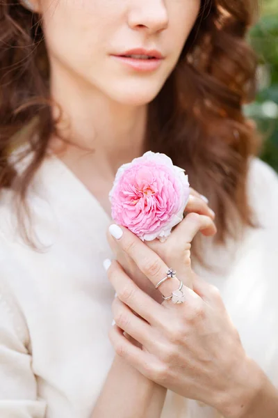 thin graceful female hands with light manicure, rings, a bracelet and a pink rose flower between fingers and part of face beautiful young woman