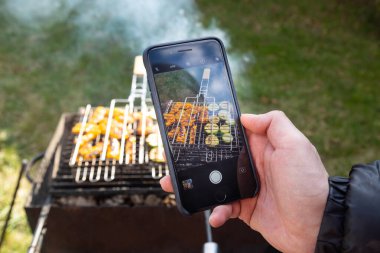 Erkek eli siyah bir cep telefonu tutuyor ve ızgara sebzelerin fotoğrafını çekiyor. Arka plandaki çimlerde dumanlı barbekü.