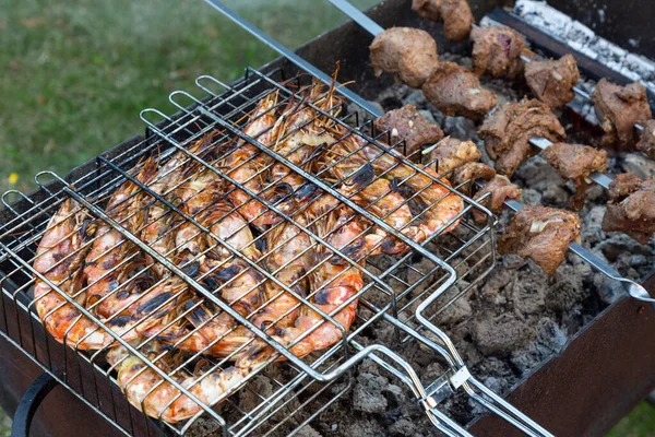 Grandes camarões cor-de-rosa langoustines com carapaça encontram-se na grelha e pedaços de carne amarrados em espetos no churrasco ao ar livre. hora de verão . — Fotografia de Stock