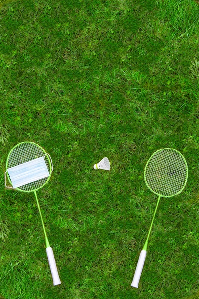 コロナウイルス対策歌 バドミントンのラケットと緑の芝生の上でシャトルチェック 1つのラケットの医療用マスク 上からの眺め — ストック写真