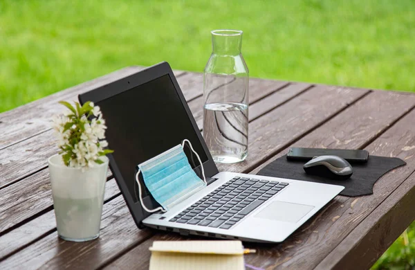 remote work or distant study from home during the coronavirus pandemic. Medical mask, laptop, mouse, notepad, mobile phone on a brown wooden table outdoor. green grass