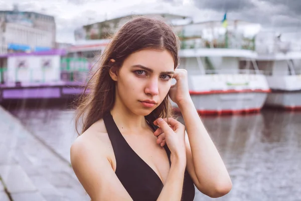 Modelo - linda morena con ojos verdes, con un vestido. Niña de pie en la lluvia sobre un fondo del río / edificios / yates. El cielo está cubierto de nubes / tormenta / tormenta nube . — Foto de Stock