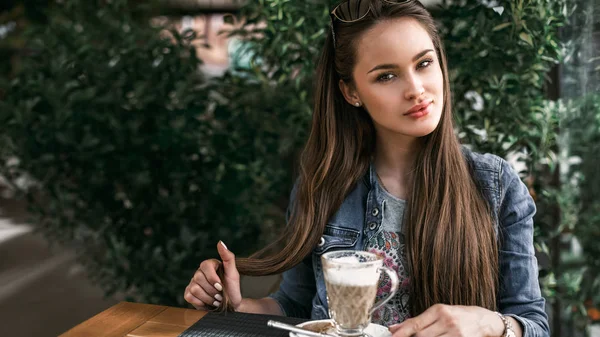 Девушка смотрит в камеру, держа волосы. Милая девушка, сидящая в кафе рядом с чашкой кофе. Горизонтальная рамка 16: 9 . — стоковое фото