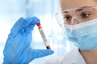 Koruyucu iş elbisesi içinde genç bir kadın tıbbi laboratuarda COVID-19 üzerinde kan örneği tüpü test ediyor.