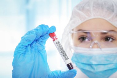 Koruyucu iş elbisesi içinde genç bir kadın laboratuarda kan örneği tüpü test ediyor.
