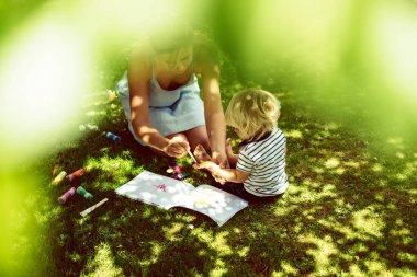 cute blonde boy drawing with young mother on green grass outdoor clipart