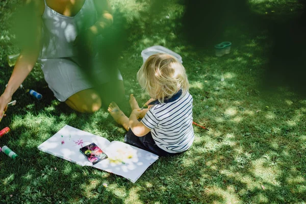 cute blonde boy drawing with young mother on green grass outdoor