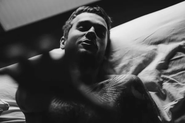 Татуированный мужчина в постели тянется к камере — стоковое фото