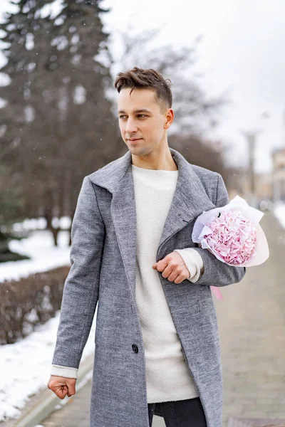 Boy friend with a bouquet of pink flowers hydrangea waiting for his girl friend outdoors while snow is falling. Valetnine`s day concept, wedding proposal.