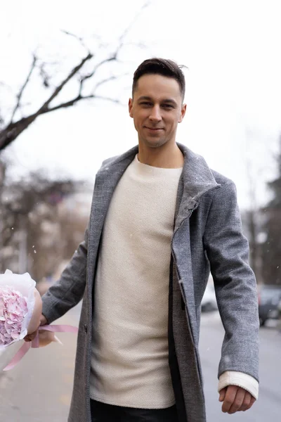 Boy friend with a bouquet of pink flowers hydrangea waiting for his girl friend outdoors while snow is falling. Valetnine`s day concept, wedding proposal.