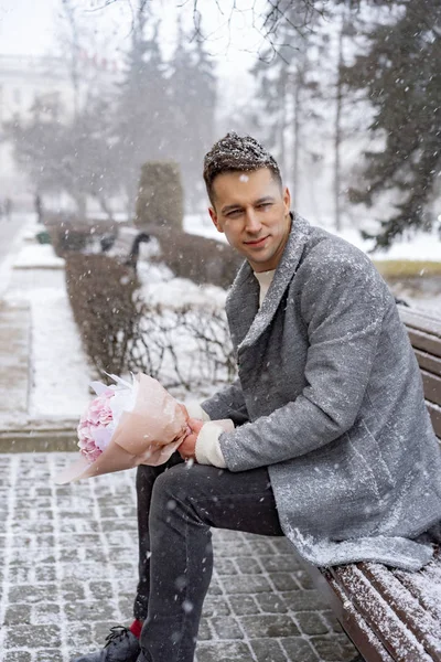 Boy friend with a bouquet of pink flowers hydrangea waiting for his girl friend outdoors while snow is falling. Valetnine`s day concept, wedding proposal.