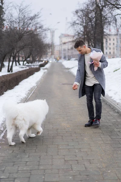 Boy friend with a bouquet of pink flowers hydrangea waiting for his girl friend and walking and playing with a dog. outdoors while snow is falling. Valetnine`s day concept, wedding proposal