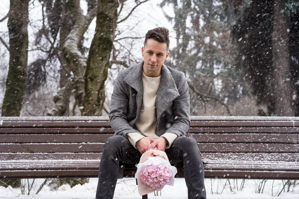 Boy friend with a bouquet of pink flowers hydrangea waiting for his girl friend outdoors while snow is falling. Valetnine`s day concept, wedding proposal.
