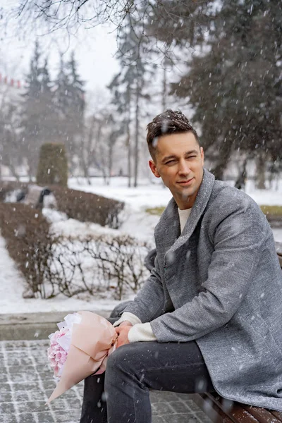 Boy friend with a bouquet of pink flowers hydrangea waiting for his girl friend outdoors while snow is falling. Valetnine`s day concept, wedding proposal.