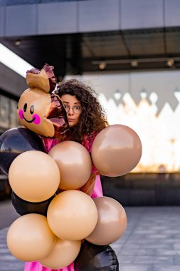 Genç, kahverengi kıvırcık saçlı, pembe uzun elbiseli, sarı, altın ve siyah balonlu bir kadın doğum günü partisine gitmek üzere sokakta yürüyor.