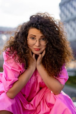 Pembe uzun elbiseli, gözlüklü, güzel, kahverengi kıvırcık saçlı kadın. Portre, yakın plan, hipster.