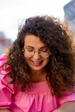 Pembe uzun elbiseli, gözlüklü, güzel, kahverengi kıvırcık saçlı kadın. Portre, yakın plan, hipster.