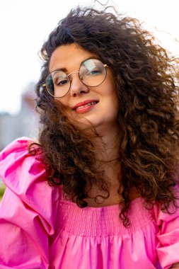 Pembe uzun elbiseli, gözlüklü, güzel, kahverengi kıvırcık saçlı kadın. Portre, yakın plan, hipster.