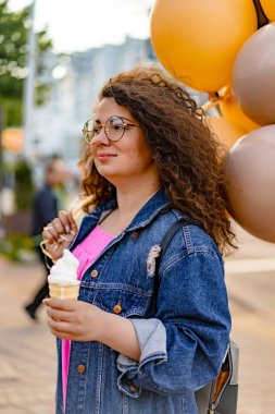 Pembe uzun elbiseli, sarı, altın ve siyah balonlu ve vanilyalı dondurma külahlı güzel, kahverengi kıvırcık saçlı kadın doğum günü partisine gitmek için caddede yürüyor.