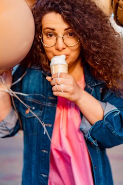 Pembe uzun elbiseli, sarı, altın ve siyah balonlu ve vanilyalı dondurma külahlı güzel, kahverengi kıvırcık saçlı kadın doğum günü partisine gitmek için caddede yürüyor.