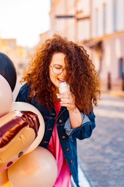 Pembe uzun elbiseli, sarı, altın ve siyah balonlu ve vanilyalı dondurma külahlı güzel, kahverengi kıvırcık saçlı kadın doğum günü partisine gitmek için caddede yürüyor.