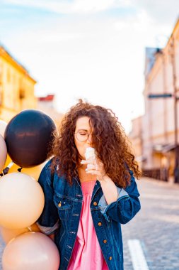 Pembe uzun elbiseli, sarı, altın ve siyah balonlu ve vanilyalı dondurma külahlı güzel, kahverengi kıvırcık saçlı kadın doğum günü partisine gitmek için caddede yürüyor.