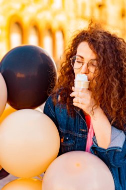 Pembe uzun elbiseli, sarı, altın ve siyah balonlu ve vanilyalı dondurma külahlı güzel, kahverengi kıvırcık saçlı kadın doğum günü partisine gitmek için caddede yürüyor.