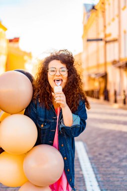 Pembe uzun elbiseli, sarı, altın ve siyah balonlu ve vanilyalı dondurma külahlı güzel, kahverengi kıvırcık saçlı kadın doğum günü partisine gitmek için caddede yürüyor.