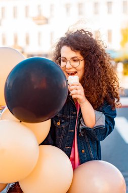 Pembe uzun elbiseli, sarı, altın ve siyah balonlu ve vanilyalı dondurma külahlı güzel, kahverengi kıvırcık saçlı kadın doğum günü partisine gitmek için caddede yürüyor.
