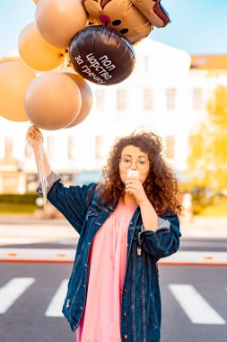 Pembe uzun elbiseli, sarı, altın ve siyah balonlu ve vanilyalı dondurma külahlı güzel, kahverengi kıvırcık saçlı kadın doğum günü partisine gitmek için caddede yürüyor.