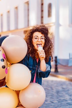 Pembe uzun elbiseli, sarı, altın ve siyah balonlu ve vanilyalı dondurma külahlı güzel, kahverengi kıvırcık saçlı kadın doğum günü partisine gitmek için caddede yürüyor.
