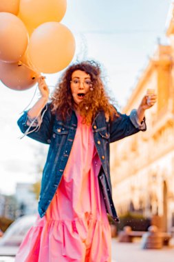 Pembe uzun elbiseli, sarı, altın ve siyah balonlu ve vanilyalı dondurma külahlı güzel, kahverengi kıvırcık saçlı kadın doğum günü partisine gitmek için caddede yürüyor.