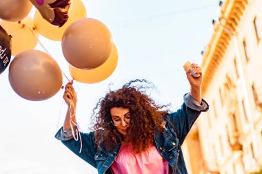 Pembe uzun elbiseli, sarı, altın ve siyah balonlu ve vanilyalı dondurma külahlı güzel, kahverengi kıvırcık saçlı kadın doğum günü partisine gitmek için caddede yürüyor.