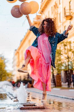 Pembe uzun elbiseli, sarı, altın ve siyah balonlu ve vanilyalı dondurma külahlı güzel, kahverengi kıvırcık saçlı kadın doğum günü partisine gitmek için caddede yürüyor.