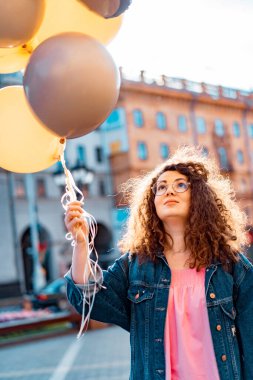 Genç, kahverengi kıvırcık saçlı, pembe uzun elbiseli, sarı, altın ve siyah balonlu bir kadın doğum günü partisine gitmek üzere sokakta yürüyor.
